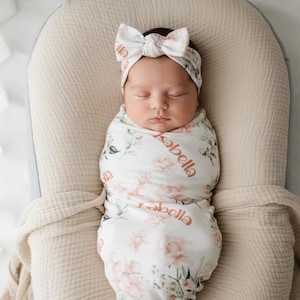 Op de afbeelding: Een pasgeboren baby gewikkeld in een bloemenprint wikkeldoek en bijpassende hoofdband. De wikkeldoek en hoofdband hebben een witte achtergrond met roze en groene bloemmotieven. De naam van de baby, "Isabella", staat op de wikkeldoek.