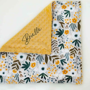 May include: A baby blanket featuring a floral design and a yellow minky dot backing. The floral pattern includes white, yellow, and green flowers and leaves on a light grey background. The name "Brielle" is written in script on the yellow side.