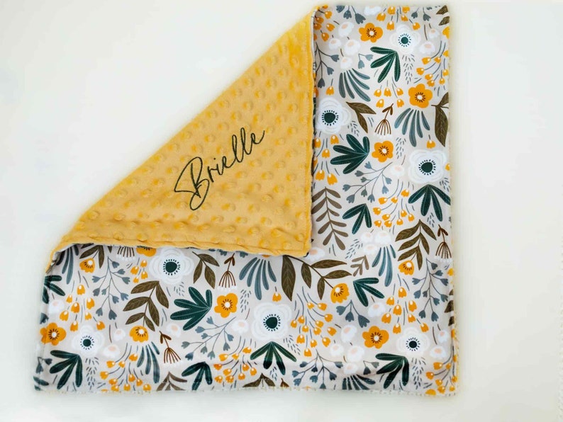 May include: A baby blanket featuring a floral design and a yellow minky dot backing. The floral pattern includes white, yellow, and green flowers and leaves on a light grey background. The name "Brielle" is written in script on the yellow side.