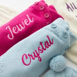 May include: Close-up of two knitted hot water bottle covers. One is bright pink with the name "Jewel" embroidered in cursive, and the other is light blue with "Crystal" embroidered in pink. Both have button details.
