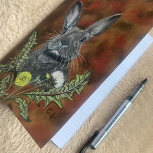 May include: A notebook with a detailed illustration of a gray rabbit with long ears, surrounded by dandelions. The cover has a brown and red background. A silver pen with a black cap rests on the lined pages.