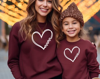 May include: Two people wearing maroon sweatshirts with white hearts and the words "mama" and "mini" inside the hearts. The sweatshirts are a matching set.