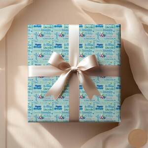 May include: A light blue gift wrap with a repeating pattern of the name "Lincoln" and the words "Happy Birthday".  The pattern also includes blue and pink balloons.  The gift wrap is tied with a beige ribbon.
