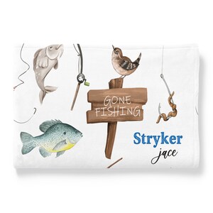 Puede incluir: Una manta de beb&eacute; blanca con tem&aacute;tica de pesca. Presenta un p&aacute;jaro marr&oacute;n, dos peces, una ca&ntilde;a de pescar, un anzuelo con un gusano y un letrero de madera que dice "Gone Fishing". La manta tambi&eacute;n tiene el nombre "Stryker Jace" escrito en texto azul.