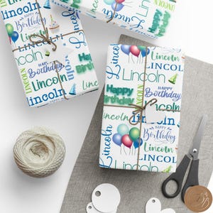 May include: White wrapping paper with a blue and green pattern featuring the name "Lincoln" and the phrase "Happy Birthday" repeated throughout. The paper is wrapped around a gift box with a brown twine bow.
