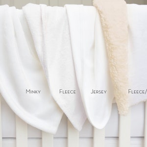 Puede incluir: Cuatro mantas blancas para beb&eacute; expuestas en una barandilla de cuna blanca. Las mantas est&aacute;n etiquetadas como "Minky", "Fleece", "Jersey" y "Fleece/Sherpa".