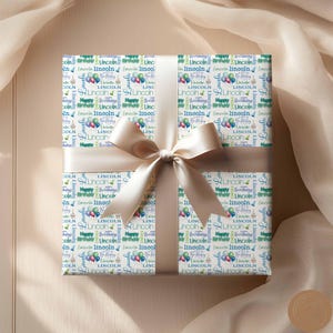 May include: A white gift wrap with a repeating pattern of blue, green, and purple balloons and the name "Lincoln" in a variety of fonts. The text "Happy Birthday" is also repeated throughout the pattern.