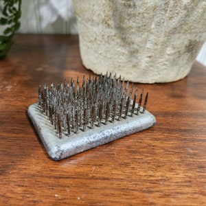 May include: A rectangular, silver-colored flower frog with numerous sharp metal pins. The pins are densely packed and protrude upwards from the base. The frog is designed to hold flower stems in place for floral arrangements. The frog sits on a wooden surface.