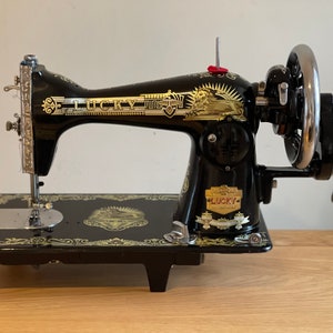 Op de afbeelding: Een zwarte vintage naaimachine met gouden accenten. De machine heeft een groot handwiel en een voetpedaal. De merknaam "LUCKY" is in gouden letters op de machine gedrukt.