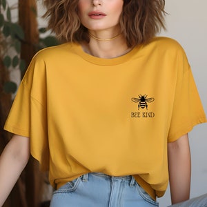 May include: Mustard yellow t-shirt with a black bee graphic and the words "BEE KIND". The shirt is a relaxed fit and is paired with blue jeans. The t-shirt is made of a soft, comfortable material.