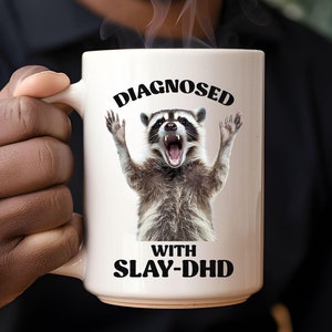 Może przedstawiać: Biały ceramiczny kubek do kawy z czarno-białym obrazkiem szop pracza z uniesionymi łapami. Na kubku nadrukowany jest tekst "DIAGNOSED WITH SLAY-DHD".