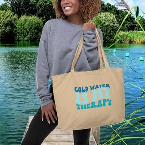 Może przedstawiać: Beżowa torba typu tote z napisem "COLD WATER IS MY THERAPY" w kolorze niebieskim i turkusowym. Torba ma długie uchwyty. Tło przedstawia jezioro i zieleń.
