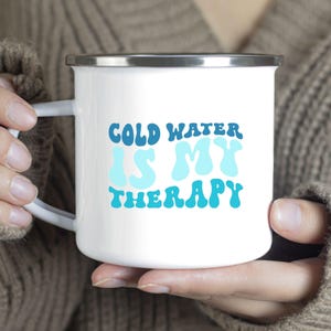 May include: White enamel camping mug with a blue and white design that says "Cold Water Is My Therapy".