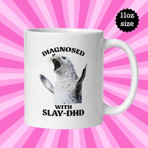 May include: White ceramic mug featuring a black and white image of a seal with the text "DIAGNOSED WITH SLAY-DHD". The mug is 11 oz.