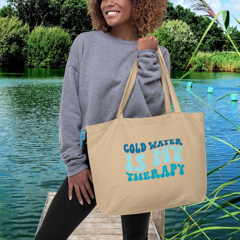 Może przedstawiać: Beżowa torba typu tote z napisem "COLD WATER IS MY THERAPY" w kolorze niebieskim i turkusowym. Torba ma długie uchwyty. Tło przedstawia jezioro i zieleń.