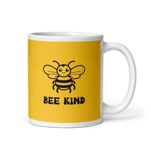 May include: White ceramic mug with a yellow background featuring a black cartoon bee and the words "BEE KIND" in a bold font. The mug has a white handle and a glossy finish.