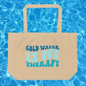 Może przedstawiać: Beżowa torba typu tote z napisem "COLD WATER IS MY THERAPY" w kolorze niebieskim i turkusowym. Torba ma prostokątny kształt z uchwytem u g&oacute;ry. Tło to niebieska powierzchnia wody.