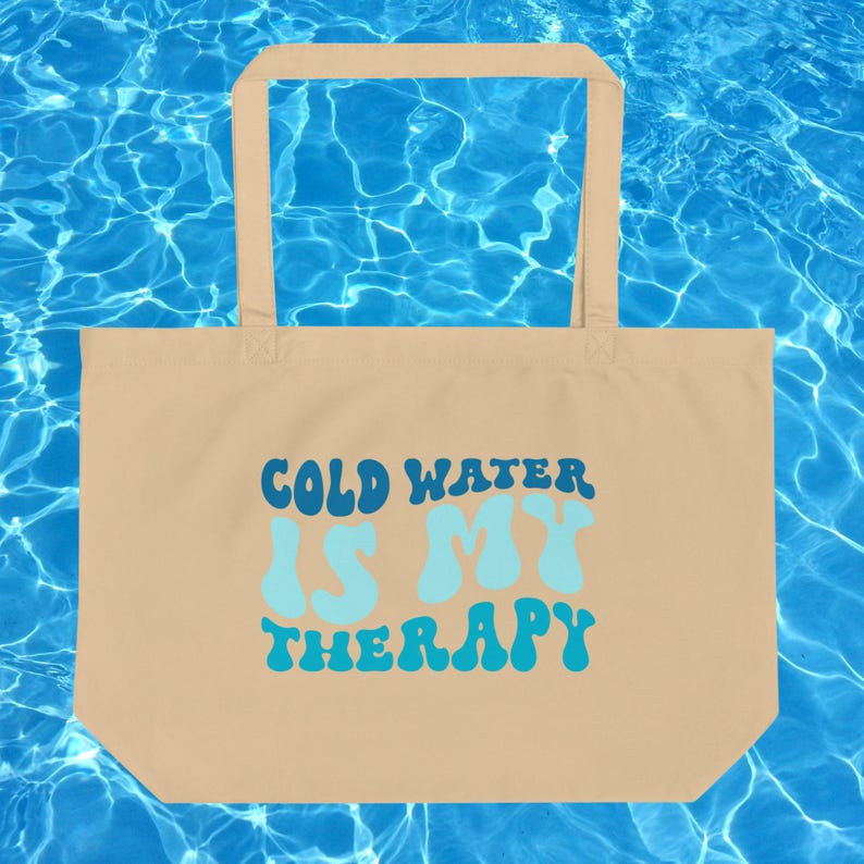 Może przedstawiać: Beżowa torba typu tote z napisem "COLD WATER IS MY THERAPY" w kolorze niebieskim i turkusowym. Torba ma prostokątny kształt z uchwytem u g&oacute;ry. Tło to niebieska powierzchnia wody.