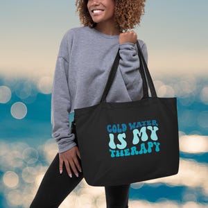 Może przedstawiać: Czarna torba typu tote z napisem "COLD WATER IS MY THERAPY" w kolorze niebieskim i jasnoniebieskim. Torba ma długie czarne uchwyty. Osoba ma na sobie szarą bluzę i czarne legginsy.