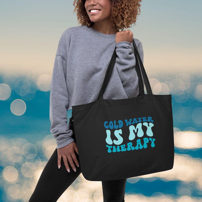 Może przedstawiać: Czarna torba typu tote z napisem "COLD WATER IS MY THERAPY" w kolorze niebieskim i jasnoniebieskim. Torba ma długie czarne uchwyty. Osoba ma na sobie szarą bluzę i czarne legginsy.