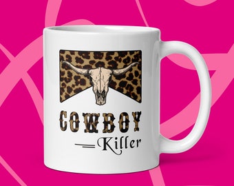 May include: White ceramic mug with a brown and black leopard print design featuring a cow skull. The text "COWBOY - Killer" is printed in black below the design.