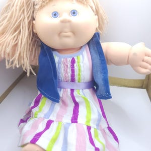 May include: A doll with light skin, blue eyes, and blonde yarn hair. The doll wears a sleeveless dress featuring vertical stripes in purple, lime green, and light blue, accented by a purple ribbon waistband and a dark blue denim-style vest.