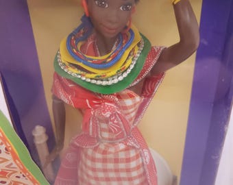 Puede incluir: Una muñeca negra que lleva un vestido de cuadros rojos y blancos, una bufanda roja y una variedad de collares coloridos. La muñeca tiene ojos marrones, cabello negro y lleva una diadema amarilla. La muñeca está en una caja con un fondo azul.