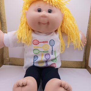 Puede incluir: Una muñeca Cabbage Patch Kid con pelo de lana amarillo que lleva una camiseta blanca con un diseño de raquetas de tenis arcoíris y pantalones negros.