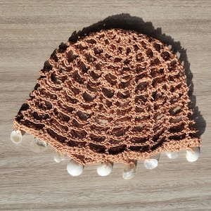 Puede incluir: Un sombrero de crochet con un diseño de red marrón y detalles de conchas blancas.