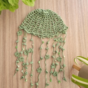 May include: A green crocheted hat with beaded fringe. The hat is made with a delicate crochet stitch and has a soft, airy feel.
