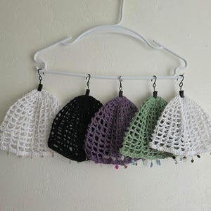 May include: Five crochet beanies in white, black, purple, sage green, and white, each featuring an open-knit mesh pattern and small dangling beads along the bottom edge. The beanies are suspended from a white plastic clothes hanger by black hooks.