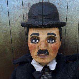 Puede incluir: Una marioneta de madera tallada a mano de Charlie Chaplin, con un bombín negro, un bigote negro y un traje negro. La marioneta tiene rasgos faciales pintados, incluyendo ojos azules y mejillas rosadas. La marioneta está suspendida por cuerdas.