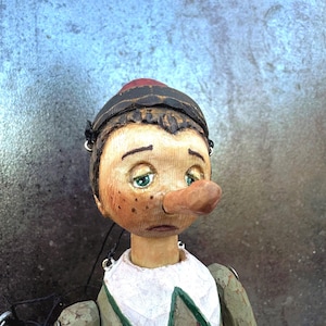 May include: A wooden puppet with a long nose, green eyes, and freckles. The puppet wears a black cap, a white collar with a red and green trim, and a green jacket. The puppet's face has a sad expression.