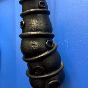 May include: A matte black, vertically oriented, cylindrical object with a textured surface. It features multiple stacked, rounded segments separated by raised horizontal rings and small, circular, protruding openings along its length, suspended by a tan twine cord.