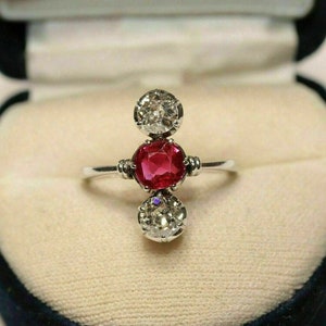 May include: A silver ring featuring a central, round, vibrant red gemstone, flanked by two clear, sparkling, round gemstones. The ring is set against a cream-colored background within a dark blue jewelry box, showcasing the ring's elegant design.
