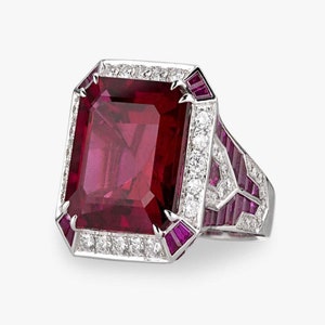 May include: A ring featuring a large, deep red gemstone surrounded by a halo of smaller diamonds and pink gemstones. The band is set with more pink gemstones.