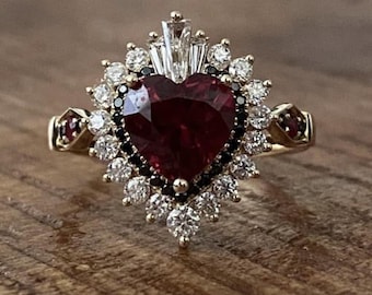 Könnte beinhalten: Ein goldener Ring mit einem herzförmigen, tiefroten Edelstein in der Mitte, umgeben von weißen und schwarzen Steinen. Zusätzliche rote Steine ​​sind auf den Schultern des Rings gesetzt, was ein Vintage-Design ergibt.