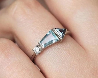 May include: A silver ring featuring a light blue, geometric-cut gemstone with a smaller, dark blue gemstone and a small, round, clear stone. The ring is set on a thin silver band, showcasing a modern design.