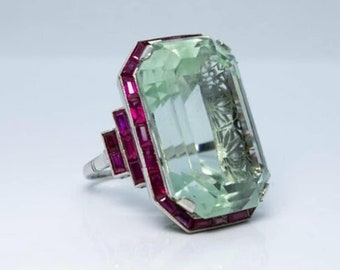 May include: A vintage ring featuring a large, light green emerald-cut gemstone surrounded by a border of small, rectangular ruby stones. The ring is set in platinum.