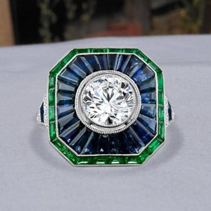 May include: A square-shaped ring with a large round diamond center stone surrounded by a halo of baguette-cut sapphires and a border of emerald-cut emeralds.