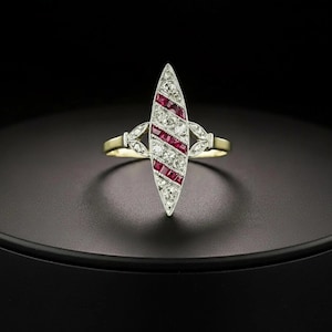 Puede incluir: Un anillo en forma de marquesa con un patrón diagonal de rubíes rojos y diamantes blancos redondos engastados en metal plateado. El anillo tiene una banda fina de color dorado y detalles metálicos en forma de hoja a ambos lados del centro.