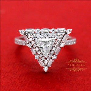 Op de afbeelding: Een diamanten ring met een driehoekig ontwerp. De ring heeft een grote, driehoekig geslepen middensteen, omringd door kleinere, baguette-geslepen en ronde diamanten. De band is ook bezet met kleine diamanten. De ring is geplaatst tegen een rode achtergrond.