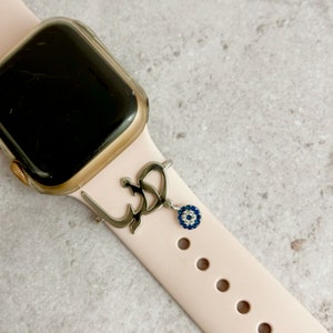 Può includere: Un cinturino rosa per Apple Watch con un ciondolo argentato. Il ciondolo è una lettera araba stilizzata e un ciondolo di occhio blu.