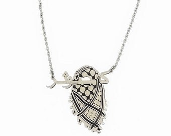 May include: A silver chain necklace with a pendant featuring a black and white patterned design of a keffiyeh, a traditional Arab headdress, with the Arabic word "Khaleeji" written above it.