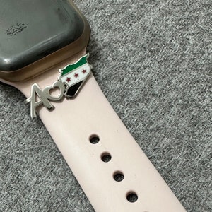 May include: A pink Apple Watch band with a silver charm attached. The charm is a heart with the letter A and a map of Syria with the Syrian flag.