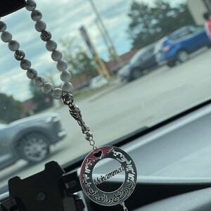 May include: A silver car charm with a heart and the name "Mohammed" inscribed in Arabic script. The charm is attached to a white beaded rosary.