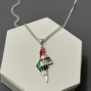 Peut inclure: Un collier en chaîne argentée avec un pendentif en forme de contour de la Palestine. Le pendentif est décoré d'un motif émaillé représentant le Dôme du Rocher, une mosquée à Jérusalem, avec des couleurs rouge, verte et blanche.