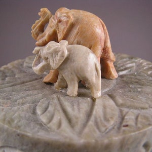 Op de afbeelding: Een gegraveerde stenen doos met een deksel met twee olifanten. De grotere olifant is bruin en de kleinere is wit.