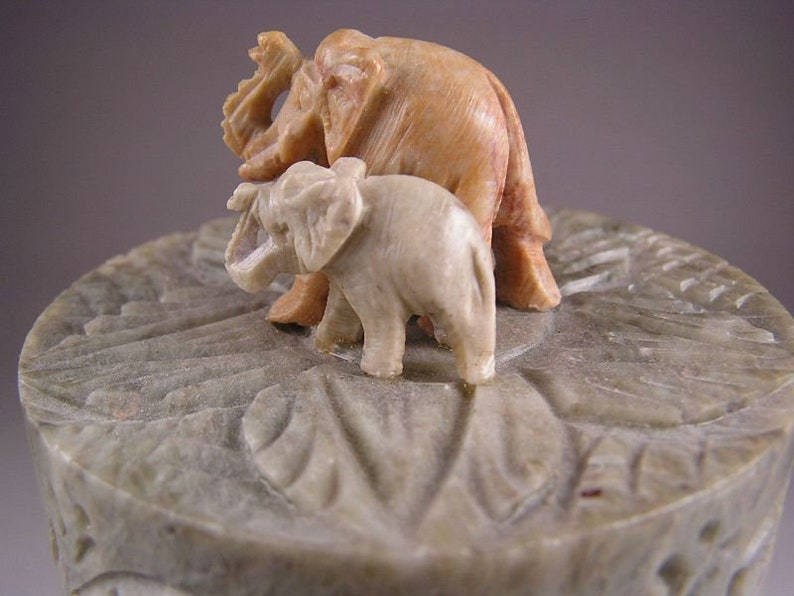 Op de afbeelding: Een gegraveerde stenen doos met een deksel met twee olifanten. De grotere olifant is bruin en de kleinere is wit.