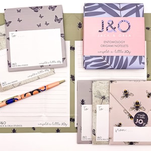 May include: A set of origami notelets with a pink and white envelope and a grey and white envelope. The notelets are decorated with a butterfly and dragonfly pattern. The envelopes are decorated with a bee pattern. The notelets and envelopes are packaged in a box with a pink and white label that says "J&O Jack & Orla Entomology Origami Notelets unfold a little joy".